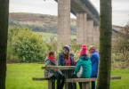 Man, woman, young girl and boy sit together at picnic bench, Boden Boo woodland, Renfrewshire Woods, underneath the Erskine Bridge