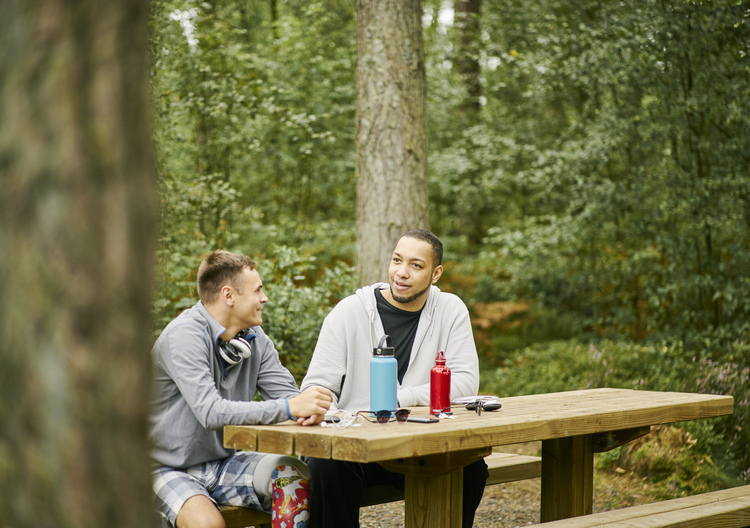 Two men (one with prosthetic leg) sit together chatting at picnic table, on woodland trail, Devilla Forest, near Kincardine