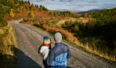 A woman in a rainbow hat and a man in a blue jacket lean against a vehicle on a forest road looking at the wooded valley below