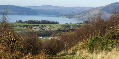 View down grassy track surrounded by scrub to green fields, open water and distant hills beyond on a bright, clear day.