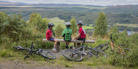 Three young boy cyclists sit on wooden bench high up in Balnain, with views overlooking Loch Ness and hills beyond