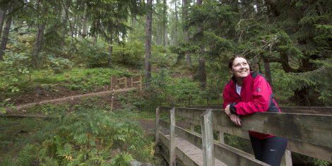 Woman in red jacket leans on wooden bridge, surrounded by trees, Cardrona Forest, near Peebles