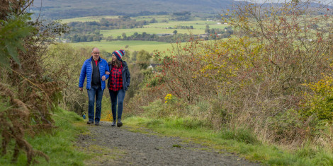 Man and woman walking uphill ona  gravel path surrounded by wild hedges and mix of trees and green fields beyond.