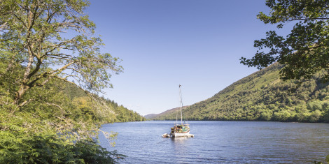 A sail boat tied up in calm water with a forested hillside in the background.