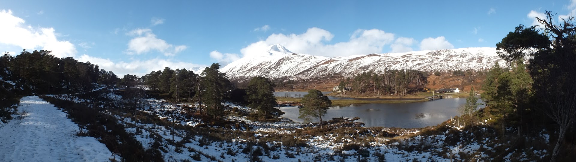Panorama of a snow-covered woodland trail with a loch and mountains in the background