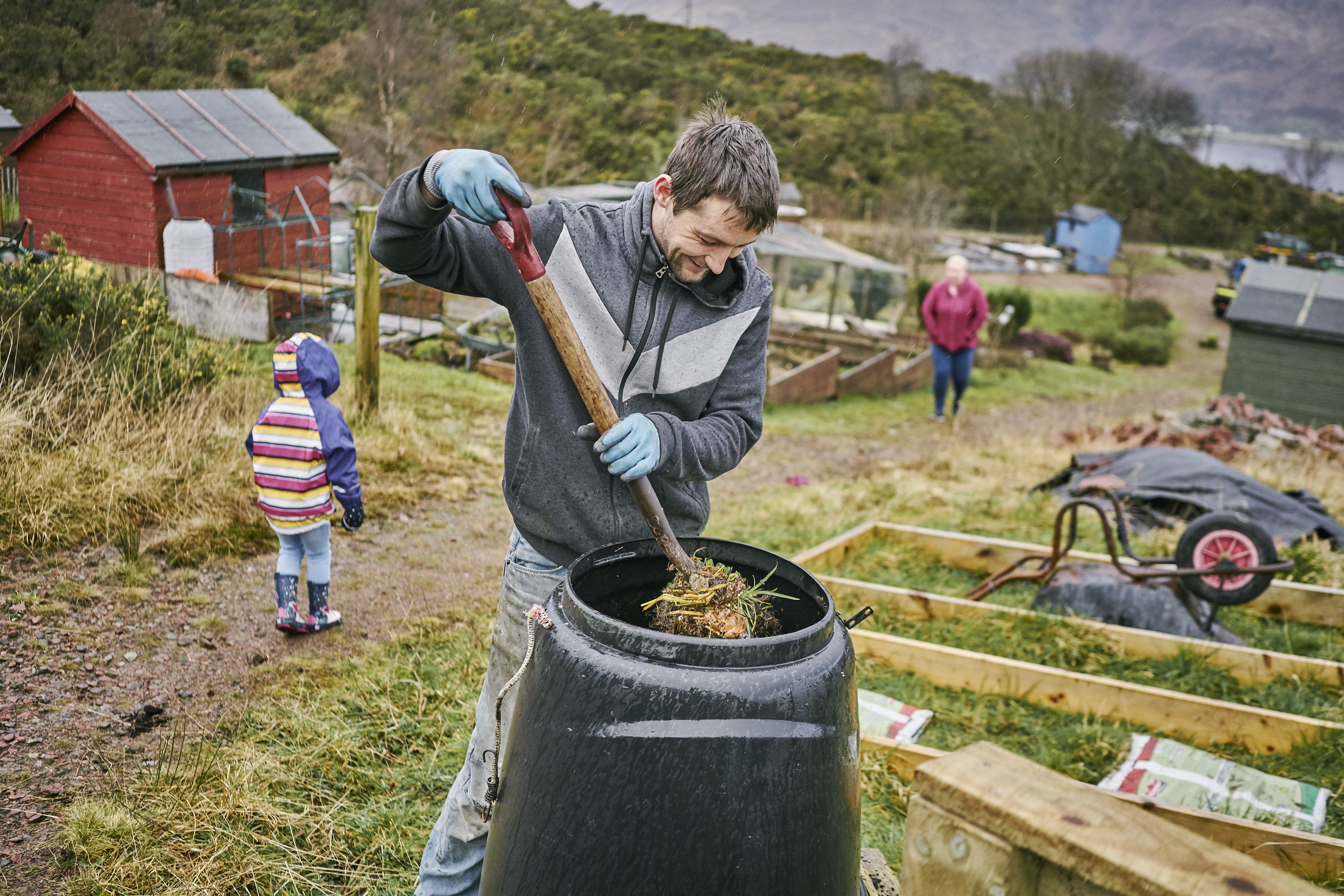 Man uses pitch fork to turn compost in plastic compost bin, with terraced raised beds and sheds of other plots behind him, as well as child and woman.