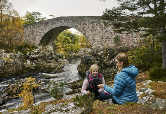 Two women and a dog next to a stone bridge and fast flowing river