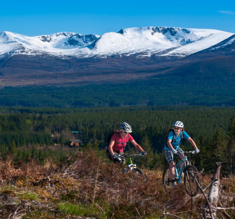 Two cyclists on a trail with vast landscape and snow covered peaks behind