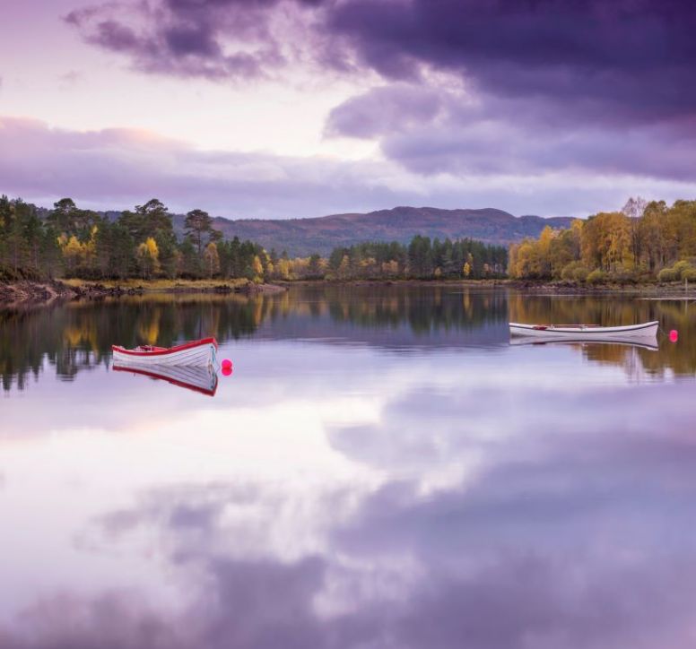 Two small boats at rest on a still loch surrounded by trees and a pink sky