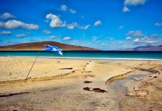 Panorama of a beach under a bright blue sky; a saltire on a flagpole is flapping in the wind.