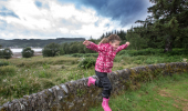 Young girl in pink jacket and wellington boots jumps from stone wall, with loch in background, Ardcastle Forest