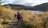 Two women and dog walking up a stony path with view beyond to loch and far wooded hillside.