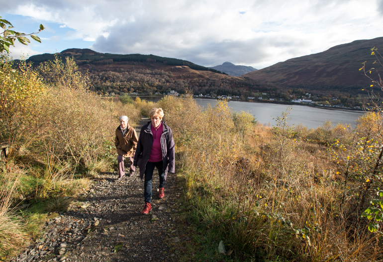 Two women and dog walking up a stony path with view beyond to loch and far wooded hillside.