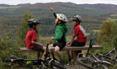  Three young boy cyclists sit on wooden bench high up in Balnain, with views overlooking Loch Ness and hills beyond