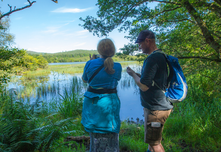 Rear view of mature man and woman looking across loch to forest on other side, Barnluasgan Oakwood Trail, Knapdale