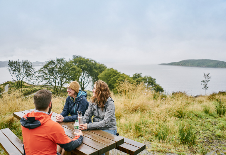 Man and two woman sit at picnic bench with views of Ardmucknish Bay in background, near Beinn Lora, Benderloch
