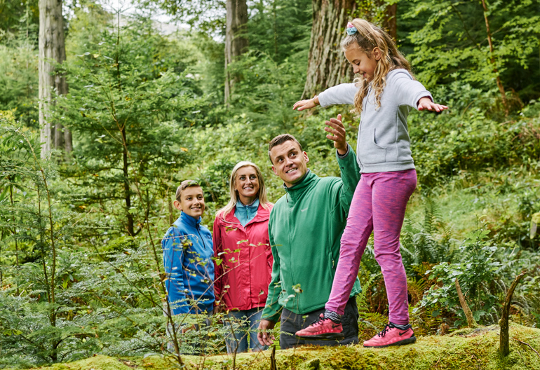 Young girl balances on moss covered fallen tree trunk, helped by man and watched on by woman and young boy, Benmore Forest, near Dunoon