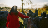 Rear view of woman in evening sun, wearing red jacket, touching boulders in Touchstone Maze, labyrinth of Scottish stones, Blackmuir Wood, near Strathpeffer