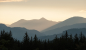 Silhouettes of distant hills, mountains and conifer forest beneath a sunny and cloudy evening sky from Blackmuir Wood