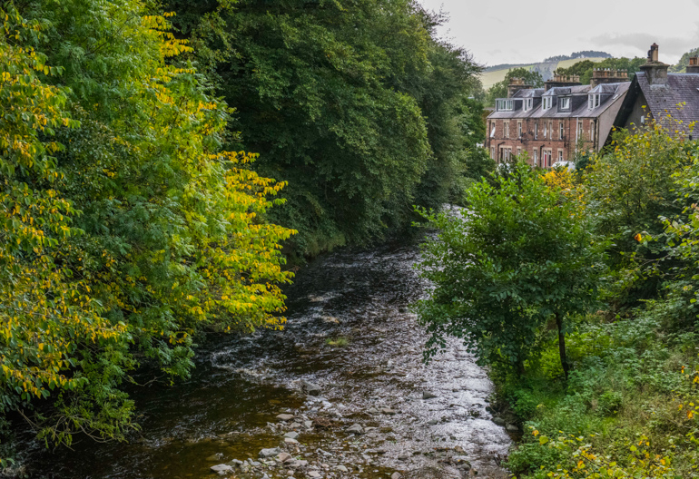 Flowing river flanked by trees, with stone houses in the background, Innerleithen, Caberston