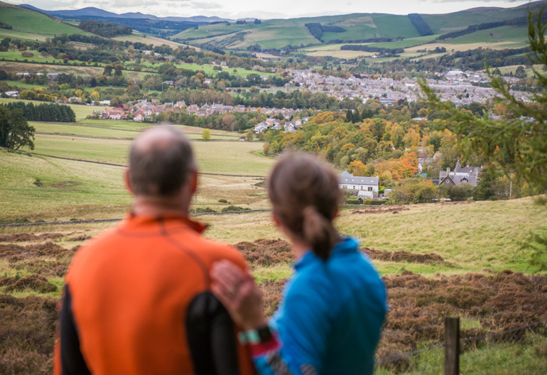 Male runner (in orange top) and female runner (in blue top) on top of hill look over towns in valley, Cademuir Forest, near Peebles