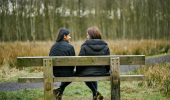 Rear view of young woman and teenage girl talking and smiling together siting on wooden bench, wearing black jackets in Cardowan Moss , Glasgow