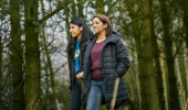 Young woman and teenage girl , in jeans and black jackets, walk arm in arm in tree lined path at Cardowan Moss, Glasgow