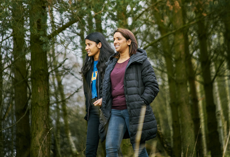 Young woman and teenage girl , in jeans and black jackets, walk arm in arm in tree lined path at Cardowan Moss, Glasgow
