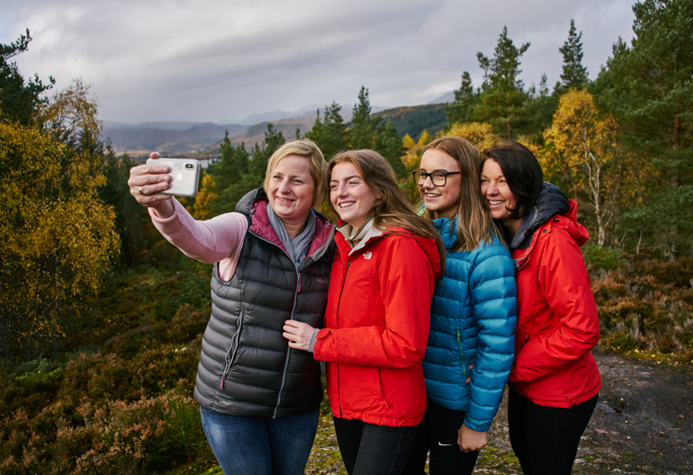 Two women and two teenage girls take a selfie photo at Contin, near Strathpeffer, with woodland and Strathconon hills in distance