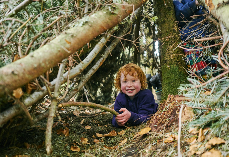 Small boy with curly ginger hair, wearing purple fleece playing with stick amidst tree branches, Craigivinean Forest, Dunkeld