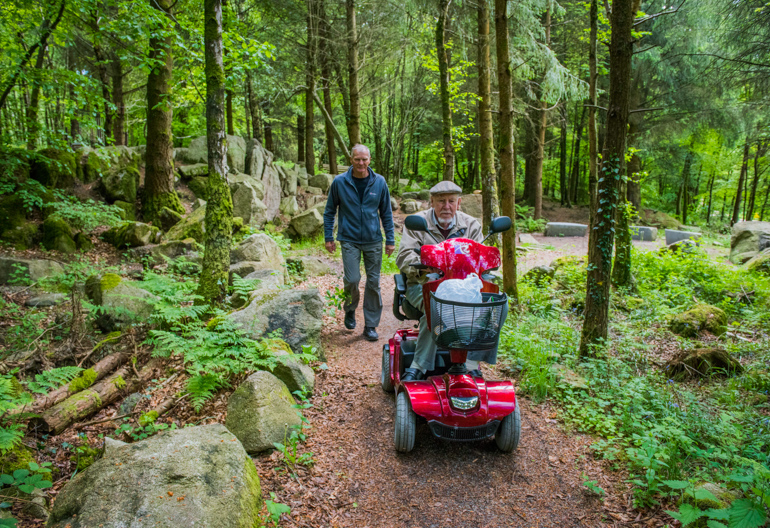 Mature man walks beside elderly man riding in mobility scooter on tree lined path, Dalbeattie Forest, near Dumfries
