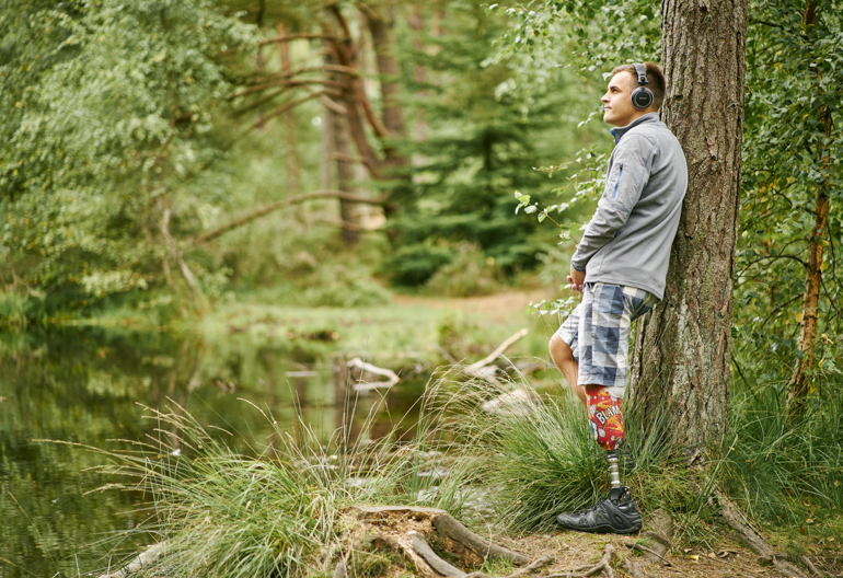  Man with prosthetic leg, wearing headphones, leans against tree on woodland trail, looking into distance, Devilla Forest, near Kincardine