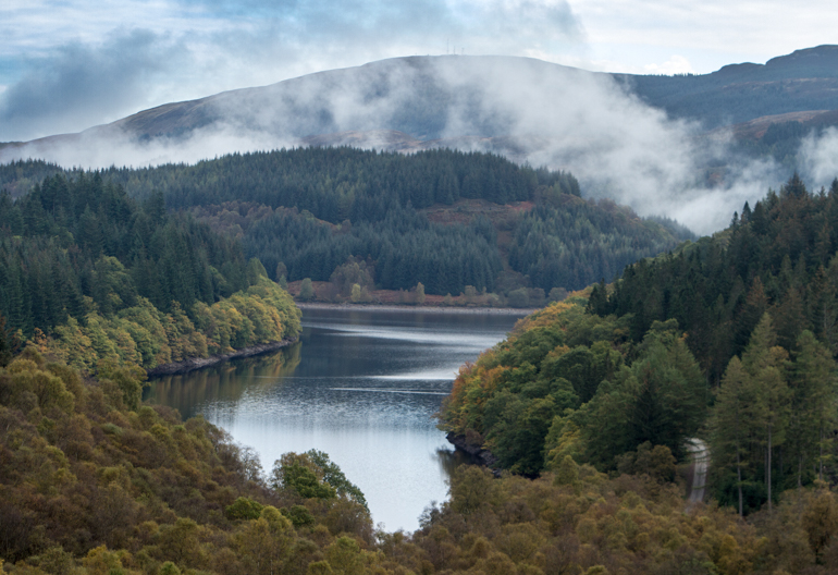  Narrow loch surrounded by heavily wooded shore with wisps of cloud rising.