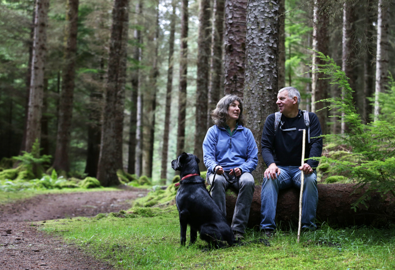  Mature man and woman walkers take a rest by sitting on fallen log under tree, with their dog, on woodland path, Farigaig forest, near Inverfarigaig
