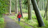 A woman walking a dog in a red rain coat along a trail by some trees.