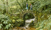 Two women walk over stone Fairy Bridge as river tumbles over rocks, Glen Creran, near Glencoe