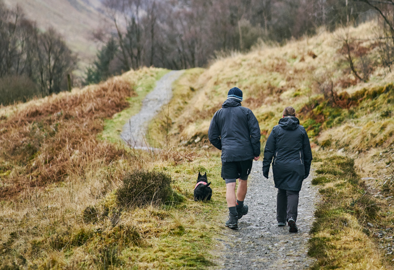 Man and woman walking along gravel path with tall trees lining each side and Scottish Terrier dog in tartan coat in front of them, Cow Hill circuit, Glen Nevis