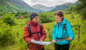 Young man and woman in walking gear inspect map together, with mountain range in background, Glen Nevis, near Fort William