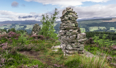 Stone carin built on the top of a hill with heather growing and a valley beyond with trees and farms