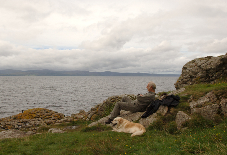 A man and dog sit on rocks looking out to sea