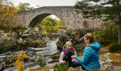 Two women pet a dog in front of a stone bridge across a rocky river