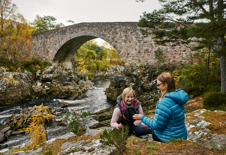 Two women pet a dog in front of a stone bridge across a rocky river