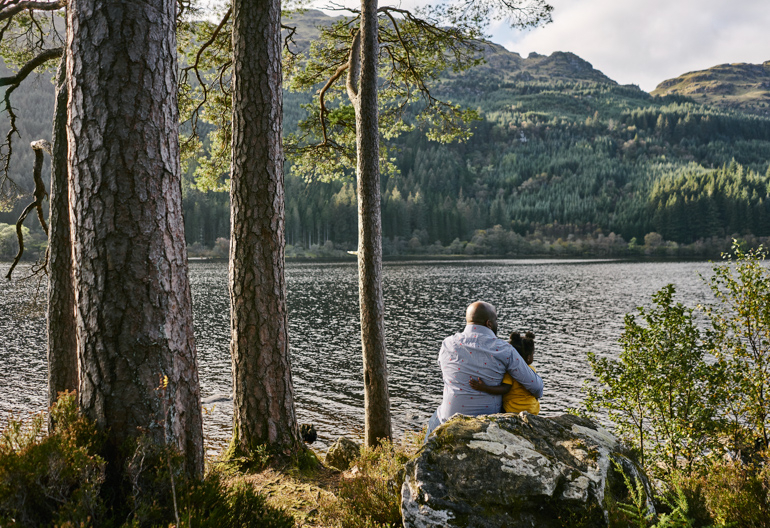 A father and daughter sit on rocks looking out over a loch