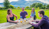 Family of four laughing while eating lunch at a picnic bench in a grassy field with loch and distant hills beyond.