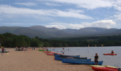 Golden sandy beach beside loch thronged with people and various canoes and kayaks, with large rounded hills beyond.
