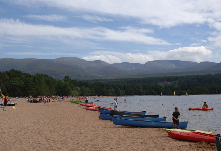 Golden sandy beach beside loch thronged with people and various canoes and kayaks, with large rounded hills beyond.