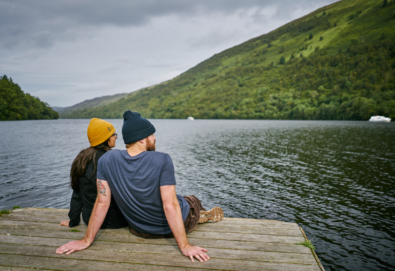 Rear view of man and woman sitting on wooden jetty, looking out to waters of Loch Oich and surrounding hills, near Fort William