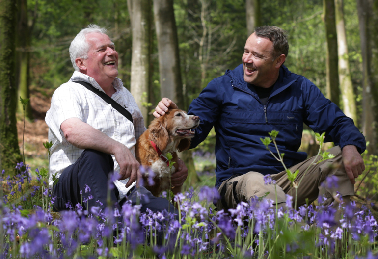 Two men and a dog witting amongst blue bells in a forest and laughing.