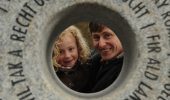 A father and daughter peak through a stone statue that has a hole cut and words cut into it