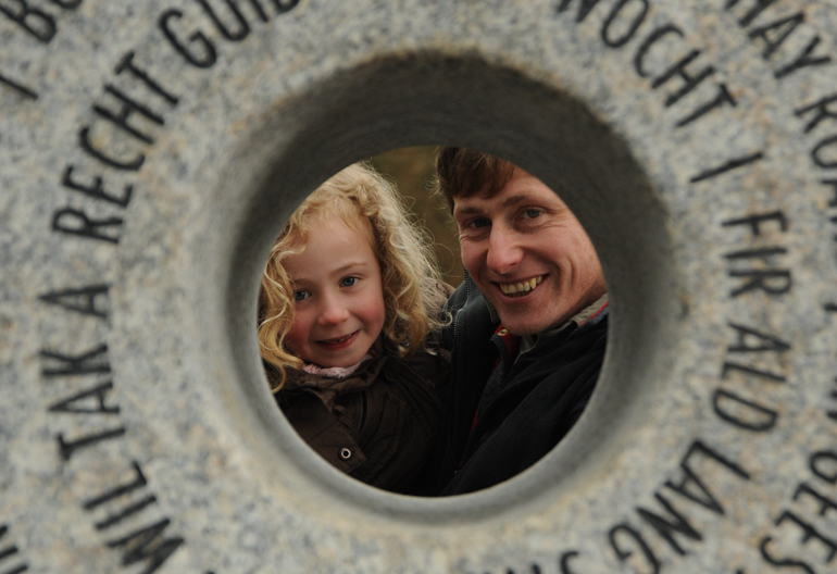 A father and daughter peak through a stone statue that has a hole cut and words cut into it
